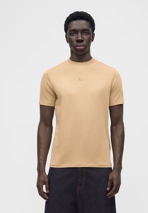 Jonge man met een donkere huid die een beige T-shirt met korte mouwen en een donkere broek draagt, staand tegen een effen lichte achtergrond.