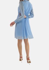 Robe bleu clair à manches longues et taille cintrée, avec un tissu transparent, une jupe fluide et des découpes sur les côtés.