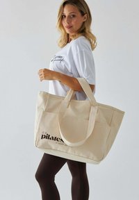 Beżowa torba na ramię z płótna z dwoma uchwytami, z napisem "jest godzina pilatesu" w pogrubionej czarnej czcionce. Prosty, zorganizowany design.
