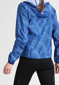 Veste de sport bleue avec un motif de vagues, en matériau léger, avec une capuche ajustée, associée à des leggings noirs avec de petites accents.