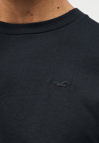 Κοντινό πλάνο ενός ατόμου που φοράει μαύρο μπλουζάκι με στρογγυλή λαιμόκοψη και μικρό, κεντητό λογότυπο πουλιού στο αριστερό μέρος του στήθους.