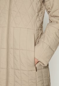 Abrigo acolchado beige con un patrón diagonal, que incluye una cremallera frontal, bolsillos laterales y una tela suave y texturizada.