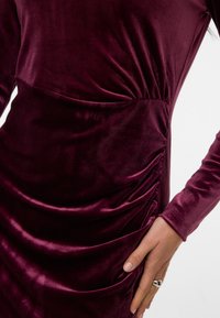 Gros plan d'une personne portant une robe en velours violet foncé à manches longues froncées, avec une bague en argent à son doigt.