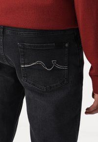 I jeans in denim scuro presentano un design semplice con una sola tasca posteriore. La tasca ha decorazioni cucite in bianco e marrone. È visibile un maglione rosso in misto lana.
