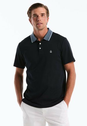 Mann mit hellbraunen Haaren, der ein schwarzes Poloshirt mit gemustertem Kragen und weißen Knöpfen sowie weiße Hose trägt, Hände in den Taschen.