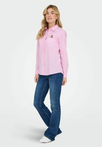 Camisa de botones de rayas rosas con un pequeño bordado de un oso en el lado izquierdo del pecho, combinada con jeans acampanados azules y zapatillas blancas.