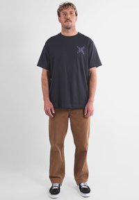 T-shirt en coton noir avec un graphique coloré sur la poitrine gauche, associé à un pantalon beige et des sneakers noirs avec des semelles blanches.