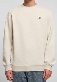Sweat-shirt beige à col rond avec manches longues, poignets et ourlet côtelés, petit logo étoile noir sur le côté gauche de la poitrine, porté avec un jean bleu.