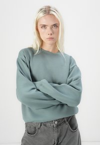 Šviesiai žalia per didelė palaidinė su apvaliu iškirpimu ir ilgomis rankovėmis, derinama su pilkomis aukštai dygsniuotomis džinsomis. Lygi tekstūra, laisvas siluetas.