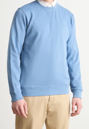 Mężczyzna w jasnoniebieskiej bluzie bez kaptura na białej koszuli z kołnierzykiem i beżowych spodniach, stojący na jednolitym jasnym tle.