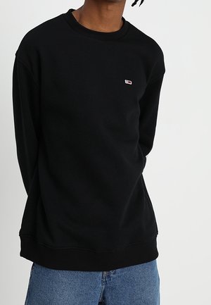 Sweat-shirt noir à manches longues et col rond avec un petit logo rouge, blanc et bleu sur la poitrine, porté avec un jean bleu, sur un fond uni.