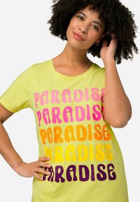 T-shirt gialla con cinque scritte "PARADISE" in colori sfumati: rosa, arancione, giallo e viola scuro. Maniche corte e vestibilità rilassata.