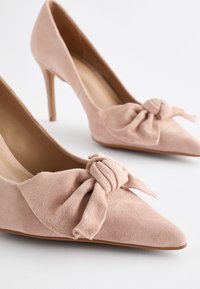 Tolmude roosa, nahast kontsaking, millel on terav varvas ja dekoratiivne sõlm ees, koos kitsase, kõrge stiletto kontsaga.
