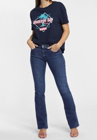 Žena oblečená v námořnickém tričku "Montego Bay", tmavomodrých džínech a pruhovaných otevřených lodičkách, stojící s rukama v kapsách na bílém pozadí.
