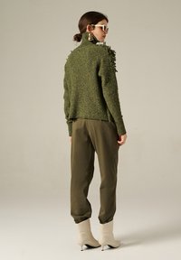 Maglione a maglia verde con texture, colletto alto e dettagli frangianti; abbinato a pantaloni olivo e stivaletti crema. La modella indossa occhiali da sole.