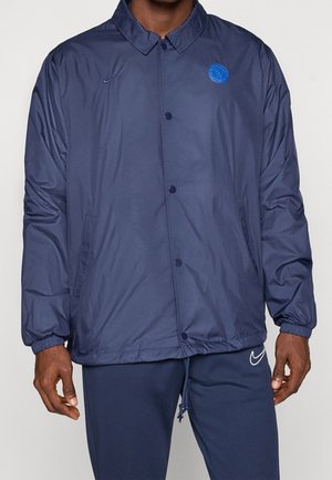 Mann trägt eine marineblaue Jacke mit Knöpfen, elastischen Bündchen, rundem Kragen, Nike-Logo und passende marineblaue Nike-Sweathosen.