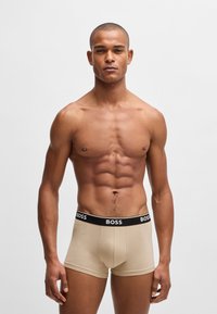Beige Boxershorts mit schwarzem elastischem Bund, der den Markennamen "BOSS" in Weiß zeigt. Glattes Material, figurbetontes Design, mittellange Beine.