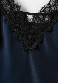 Morska bluza s detaljnim crnim čipkastim ovratnikom s cvjetnim uzorcima i izdriganim rubovima, izrađena od glatkog materijala.