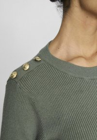 Pull vert côtelé avec un col rond, orné de trois boutons décoratifs dorés sur l'épaule, texture lisse et coupe ajustée.