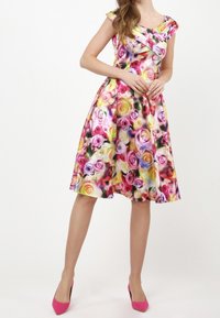 Blumenkleid mit kurzen Ärmeln, das mit einem bunten Rosenmuster in Pink, Gelb und Lila verziert ist. Eng anliegendes Oberteil, ausgestellter Rock, kombiniert mit pinken High Heels.