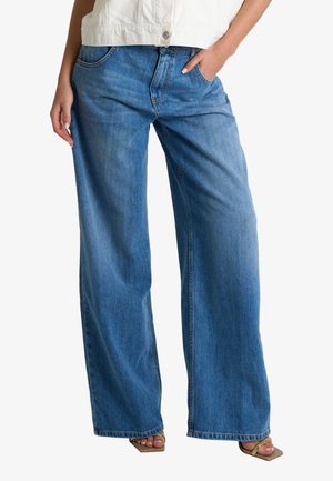 Persona che indossa jeans blu a vita alta e gamba larga e sandali aperti color cuoio, con una mano in tasca e top bianco parzialmente visibile.