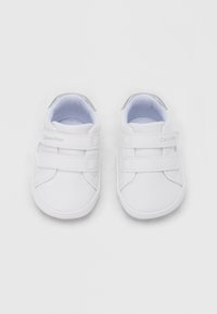 Beli otroški supergi z dvema trakovoma na ježka, gladkim sintetičnim materialom, okroglim prstom in srebrnim poudarkom na peti.