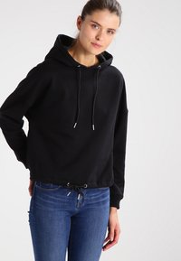 Sudadera corta negra con capucha ajustable, que presenta cordones con puntas de plata y un fondo ceñido. Combinada con jeans azules.