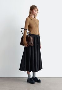 Tan ribbed long-sleeve top with button placket, black pleated maxi skirt, gray socks, and black loafers, holding a brown leather handbag.