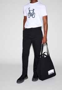 Vit t-shirt med ett blått cykelmotiv, kombinerad med svarta byxor och svarta sneakers. En svart tygväska med en vit etikett hålls i handen.