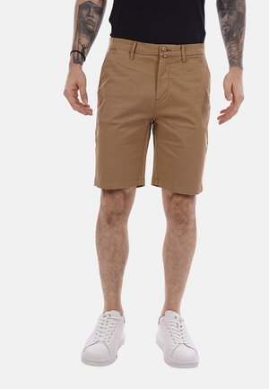 Uomo che indossa pantaloncini beige fino al ginocchio, scarpe da ginnastica bianche e una maglietta nera, mostrando gli avambracci tatuati su uno sfondo semplice.