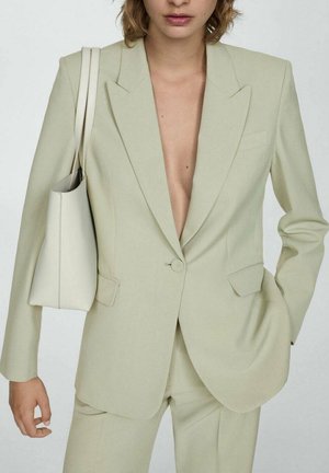 Femme portant un costume beige avec un blazer à un bouton et un pantalon assorti, portant un sac à bandoulière beige clair sur son épaule gauche.
