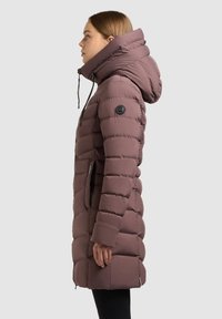 Žena u profilu nosi zimski kaput do koljena u boji mauve, podstavljen i s kapuljačom, visokim ovratnikom, protiv jednobojne pozadine.