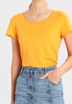 Kobieta ubrana w pomarańczowy t-shirt z krótkim rękawem, wpuszczony w wysokowydatną spódnicę dżinsową z guziczkami z przodu, z jedną ręką w kieszeni.