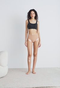 Schwarzes Crop-Top und hautfarbene High-Waist-Unterwäsche an einem Model. Glatter Stoff, enganliegendes Design, weiche Textur, stehend auf einem hellen Teppich.