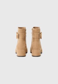 Bottines en daim tan avec un talon bloc, une fermeture éclair latérale et un accent circulaire. Texture lisse avec un design minimaliste et des lignes épurées.