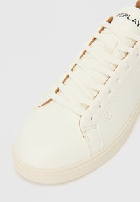 Baskets blanches en cuir avec un design perforé sur le côté et des lacets blancs plats. Présente un logo sur la languette et des accents beige clair près de l'orteil.