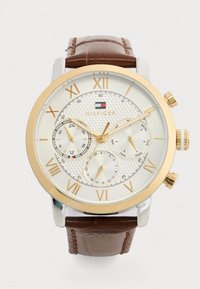 Ανδρικό ρολόι χειρός με λευκό καντράν, χρυσούς ρωμαϊκούς αριθμούς, τρεις υποκαταστάτες για 24-ωρη ώρα, ημέρα και ημερομηνία, χρυσή κάσα και καφέ δερμάτινο λουρί.