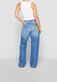 Světle modré kalhoty s širokými nohavicemi a vybledlým texturem, s dvěma zadními kapsami a žlutou nášivkou úsměvné tváře na pravé kapse.