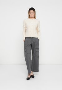 Maglione color beige chiaro a trecce abbinato a pantaloni cargo grigi larghi con molte tasche. La modella indossa scarpe nere con punta affusolata.