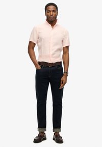 Jasnoróżowa koszula z krótkim rękawem, z guzikami, wykonana z lnu. Ciemnoniebieskie dżinsy z podwiniętymi nogawkami i brązowym skórzanym paskiem. Brązowe mokasyny.