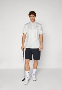 Világosszürke mintás galléros póló három gombbal és fekete Nike logóval, sötét rövidnadrággal és fehér zoknival párosítva. Golfütőt tart a kezében.