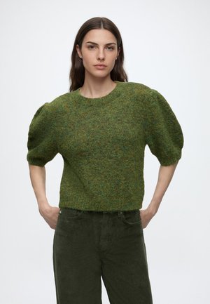 Femme portant un pull vert texturé à manches bouffantes et un pantalon vert foncé, debout, les mains sur les hanches, devant un fond uni.