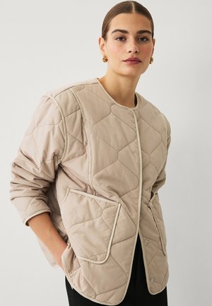 Woman with slicked-back hair wearing gold hoop earrings and a beige quilted jacket with large front pockets, standing against a plain background.