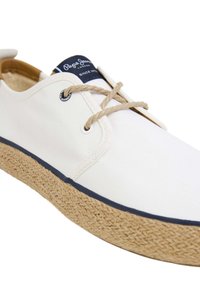 Scarpa bianca in tela con suola in juta intrecciata. Presenta lacci beige, etichetta di marca blu navy e bordatura blu scuro lungo la suola.