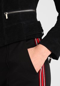 Ruka v černých kalhotách s červenými a bílými bočními pruhy, oblečená v černé bundě na zip s přezkou v oblasti pasu.