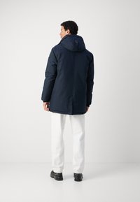 Navy blue parka with a hood, features a smooth texture, long sleeves, and a straight cut, paired with white pants and black shoes.