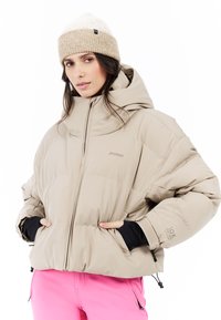 Beige pufferjack met een capuchon, ritsluiting en elastische manchetten. Het model draagt een roze broek en een lichtbeige gebreide beanie.
