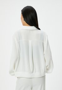 Blusa bianca in satin con una vestibilità morbida, colletto strutturato e dettagli con pieghe sul retro. Presenta polsini e orlo a coste.