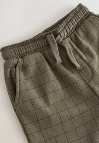 Pantaloni della tuta in cotone di colore verde oliva con motivo a griglia. Presentano un elastico in vita e un cordino per la regolazione. Texture morbida.