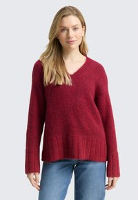 Kobieta o długich blond włosach, ubrana w czerwoną, miękką sweter typu V-neck i niebieskie dżinsy na jednolitym jasnym tle.